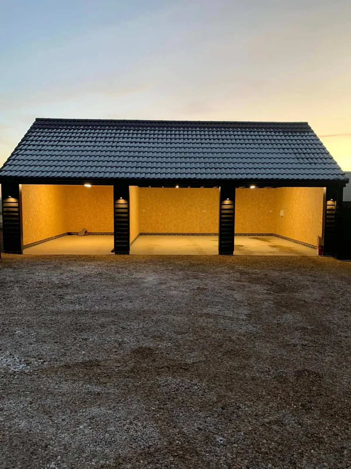 Open-fronted triple garage lit at dusk, with internal bay lighting and external lights on the posts